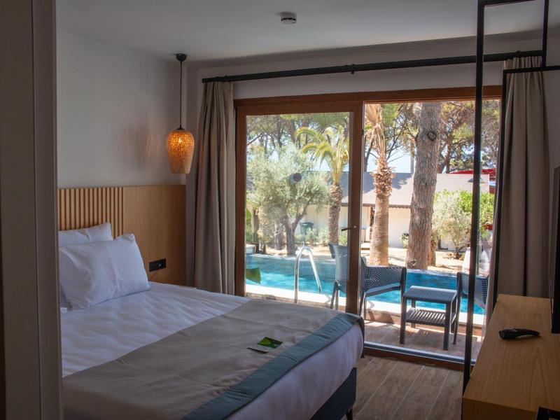 Jasny pokój hotelowy z łóżkiem podwójnym i widokiem na basen i ogród przez duże drzwi przesuwne.