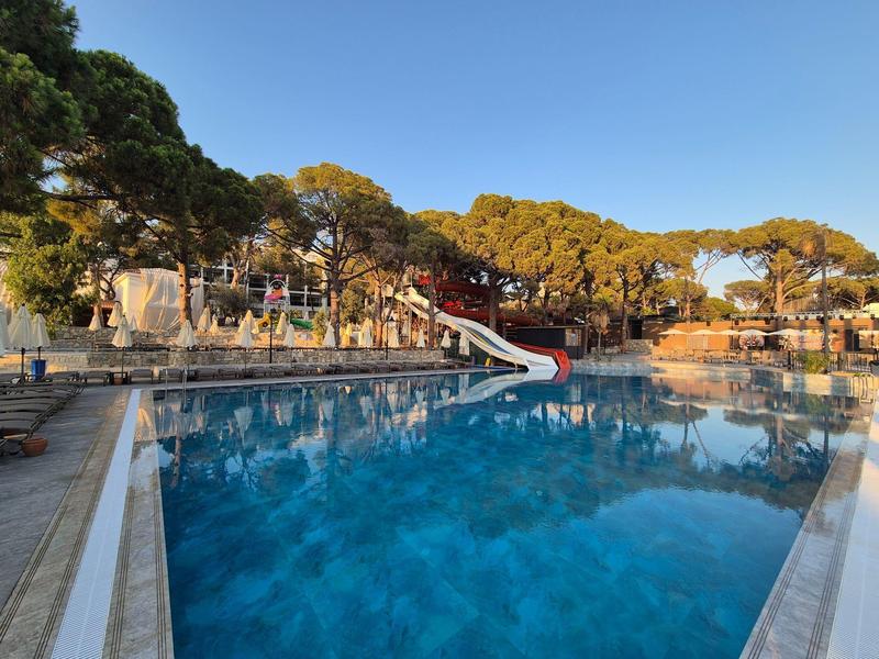 Duży basen na zewnątrz z zjeżdżalnią, otoczony drzewami i restauracjami pod czystym niebem.