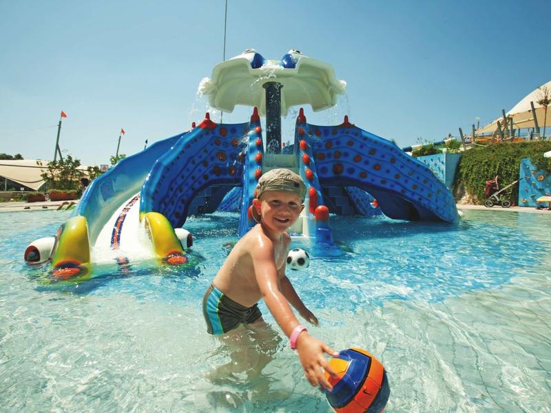 Child playing with ball in shallow water in front of octopus water slide at pool.