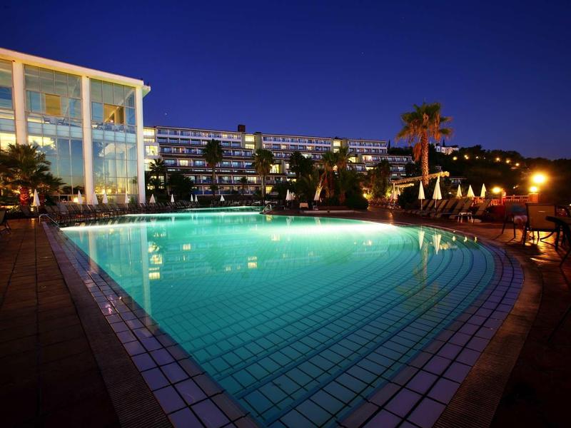 Illuminated hotel pool at night with surrounding buildings and palm trees.