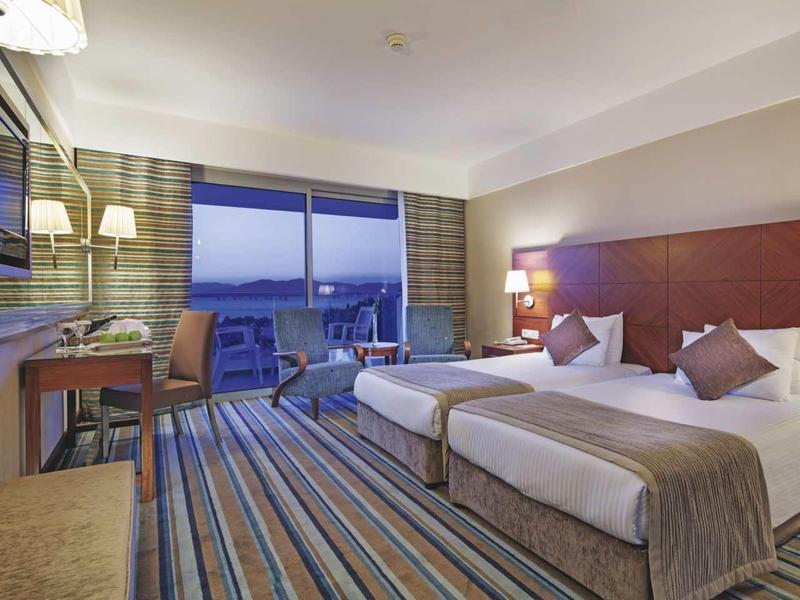 Modern hotel room with two beds, desk, and city view at dusk.