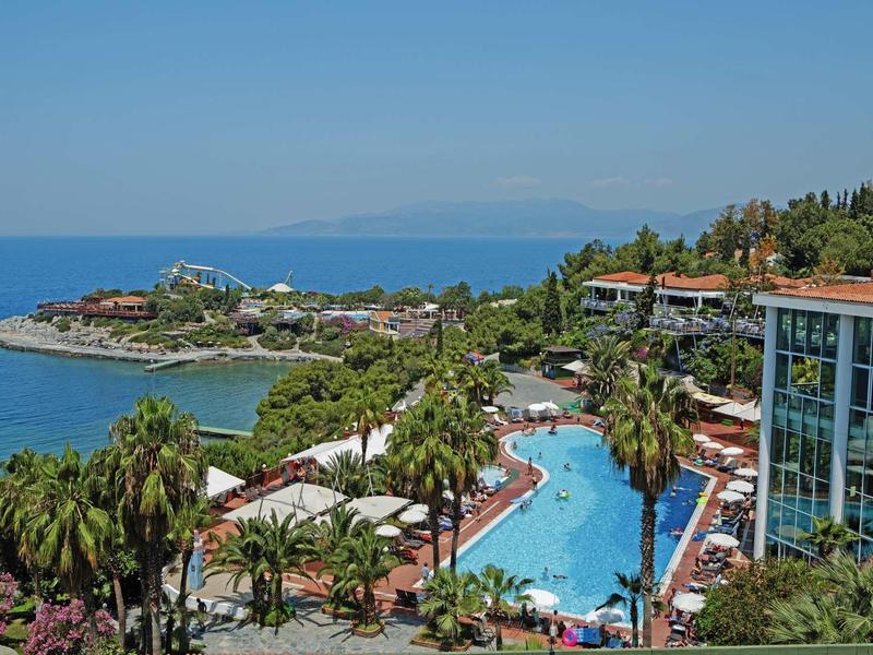 Hotel resort with large pool, palm trees, and sea view under clear sky.