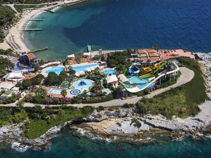 Aerial view of a small island with a hotel complex, pools, and beach by clear blue sea.