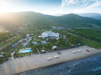 Letecký pohled na hotelový komplex s bazénem u pláže a horami v pozadí.