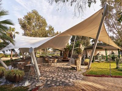 Zastíněná venkovní terasa s stolky a židlemi pod bílým plátnem, obklopená vegetací.