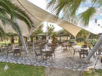 Sitzbereich im Freien unter weißen Sonnensegeln und Palmen mit Blick auf eine grüne Landschaft.