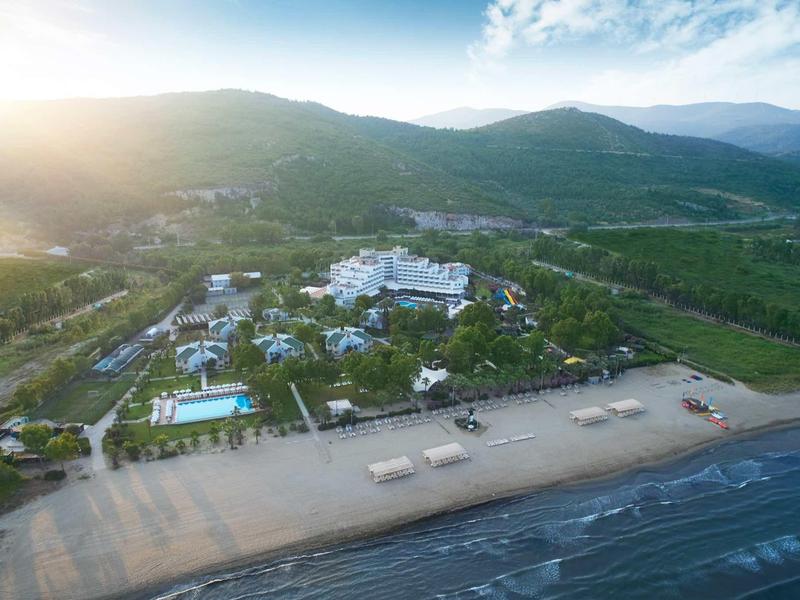 Letecký pohled na hotelový komplex s bazénem u pláže a horami v pozadí.