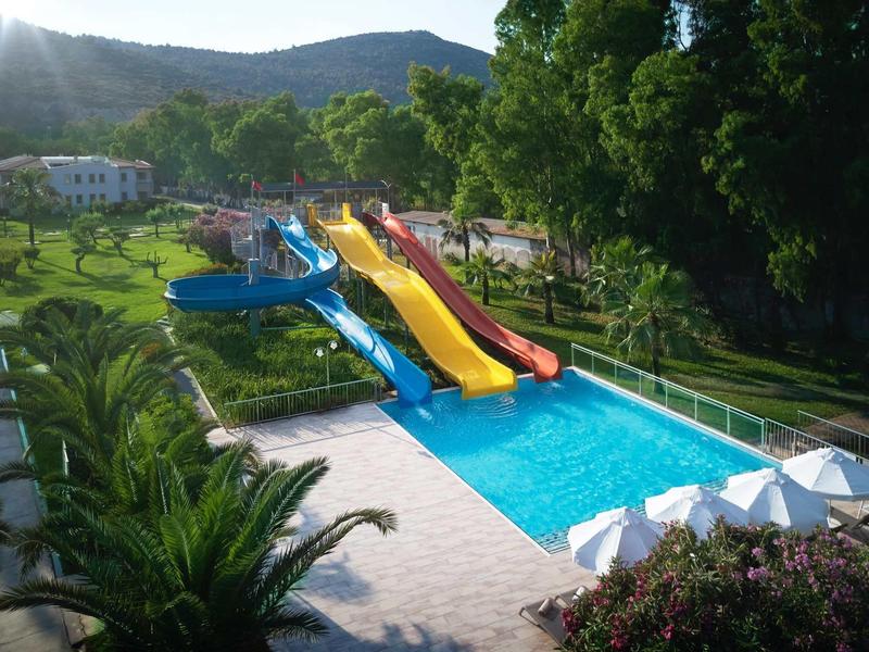 Barevné skluzavky vedoucí do bazénu obklopeného zelení a bílými slunečníky.