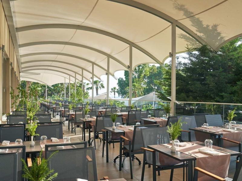 Venkovní terasa restaurace s stolky a židlemi pod velkým bílým baldachýnem.