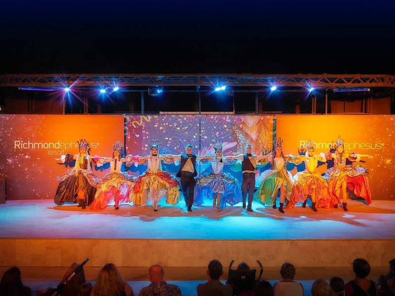 Barevné taneční vystoupení na pódiu s publikem v útulné atmosféře.