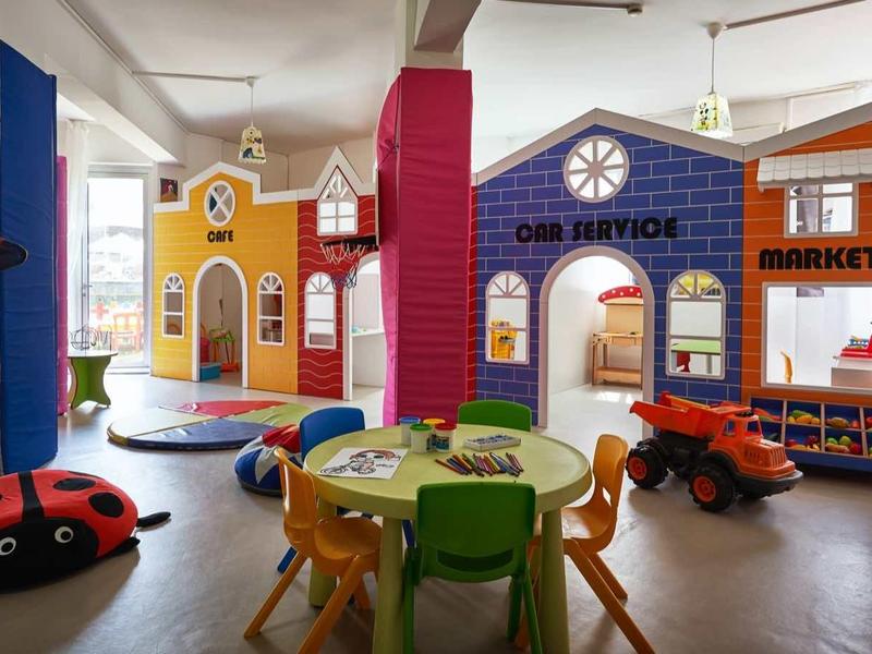 Barevná dětská herna s stolky, židlemi a domečky v miniatuře.