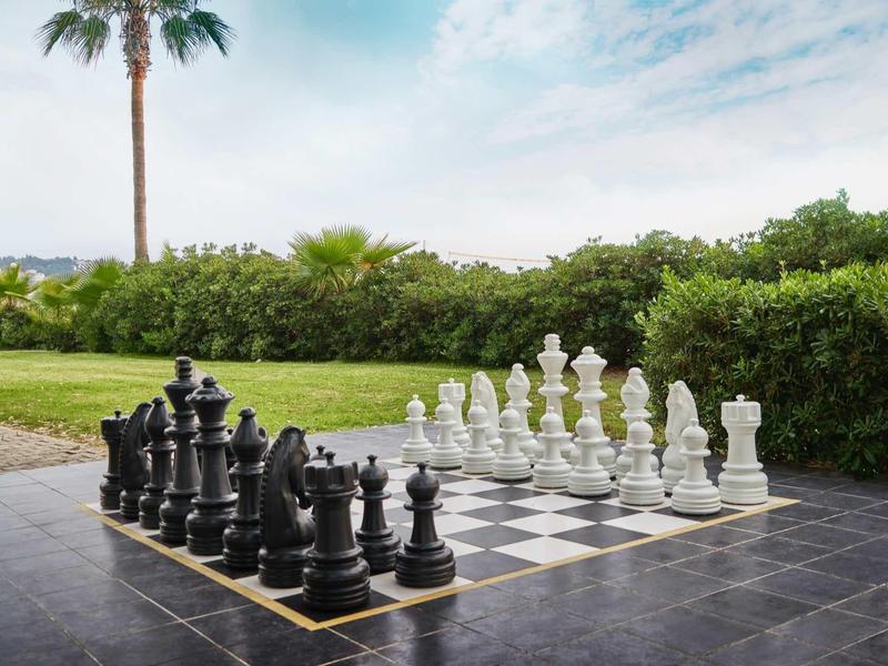 Obrovská venkovní šachovnice na terase obklopená zelení pod částečně oblačnou oblohou.