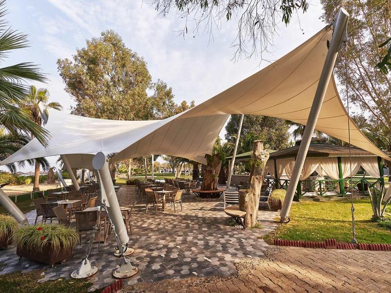 Zastíněná venkovní terasa s stolky a židlemi pod bílým plátnem, obklopená vegetací.