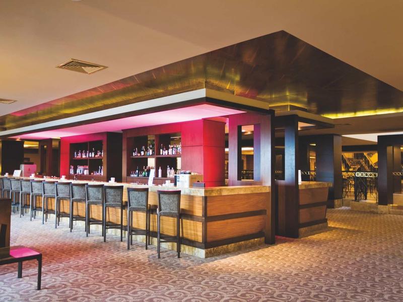 Moderne Hotelbar mit Holztheke, Lederhockern und warmem, indirektem Licht in hellem Raum.