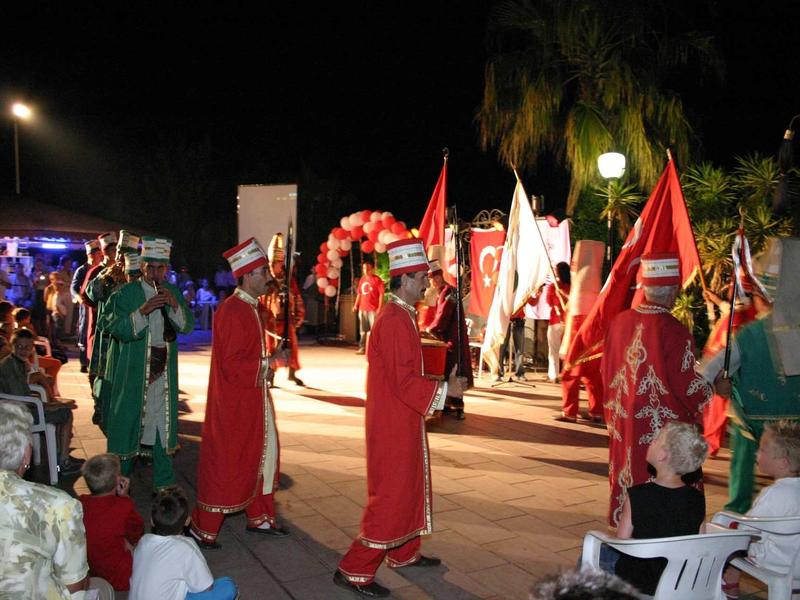 Nachtliche Prozession mit Menschen in bunten Kostümen und roten Umhängen, Fahnen und Lichtquellen sichtbar.