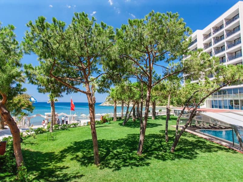 Green hotel garden with pine trees, sea view, and swimming pool under a blue sky.