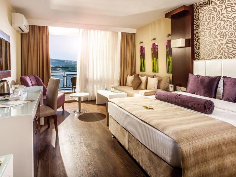 Bright hotel room with double bed, seating area, and sea view through large windows.