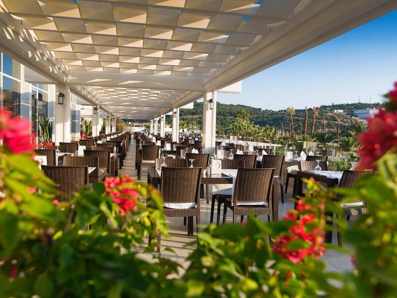 Large hotel terrace with modern tables and chairs, surrounded by colorful flowers and hills in the background.