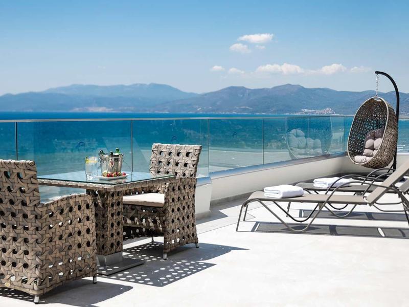 Modern terrace with wicker furniture, sun loungers, and sea view under blue sky.