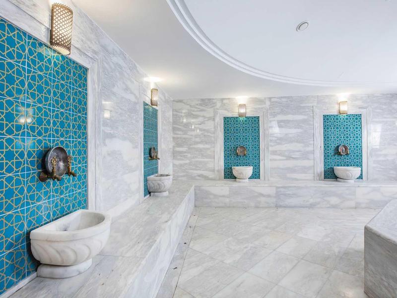Interior view of a luxurious hammam with white marble walls and blue mosaic niches.