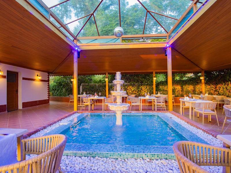 Piscina interior con fuente y área de asientos bajo un techo de vidrio con vista a árboles.