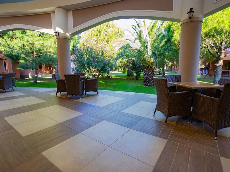 Terraza cubierta con mesas y sillas, rodeada de jardín y árboles en un complejo hotelero.