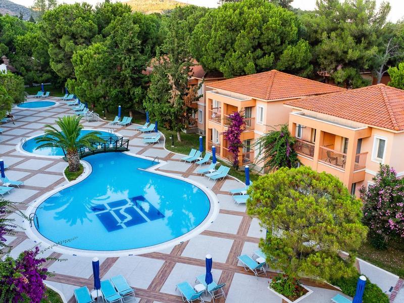 Complejo hotelero con piscina ovalada, tumbonas y rodeado de árboles verdes y edificios con techos rojos.