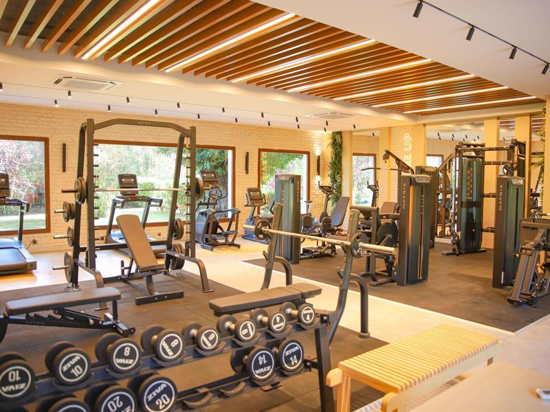 Gimnasio luminoso con varios equipos de entrenamiento y pesas frente a grandes ventanas.