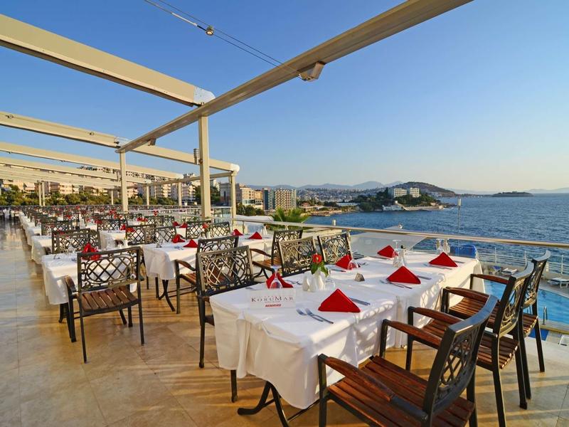 Terrassenrestaurant mit weißen Tischdecken, roten Servietten und Meerblick bei klarem Himmel.