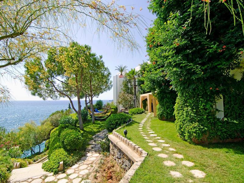 Grüner Gartenweg mit Rasen, Bäumen und Blick aufs blaue Meer unter blauem Himmel.