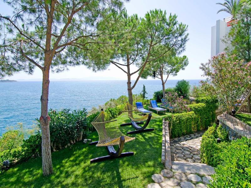 Grüner Garten mit Bänken und Bäumen am Meer, blauer Himmel, steiniger Weg führt durch Vegetation.