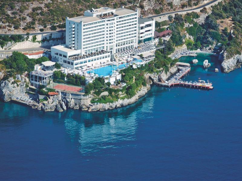 Großes weißes Hotel am Meer, umgeben von Felsen, mit Pool und Steg. Hügel im Hintergrund.