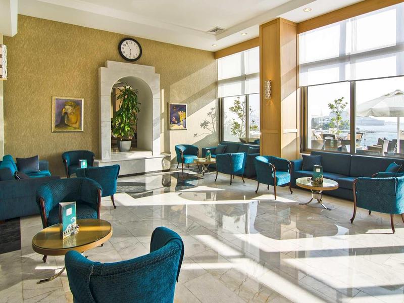 Großer heller Raum mit blauen Sesseln, gläsernen Tischen und großen Fenstern mit Stadtblick.