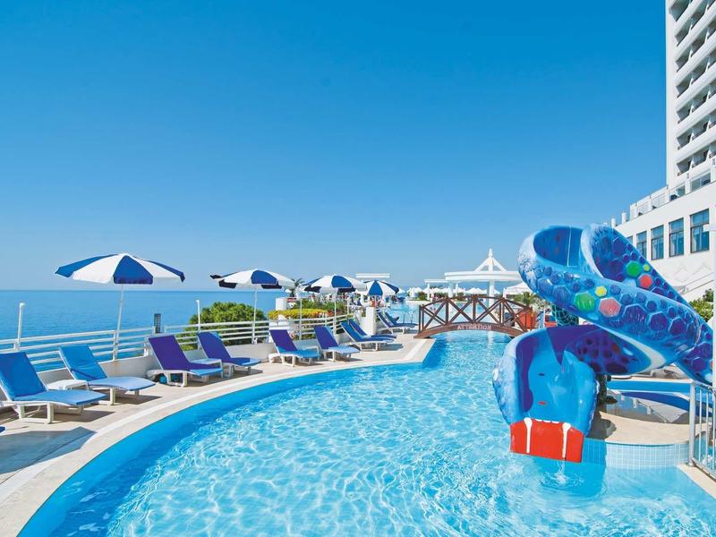 Pool mit Wasserrutsche, Liegestühlen und Sonnenschirmen neben einem hohen Hotelgebäude am Meer.