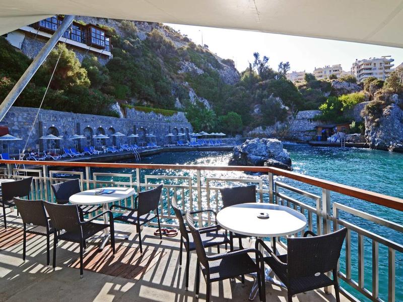 Terrassenbereich mit Tischen und Stühlen am Wasser, umgeben von Felsen und Gebäuden im Hintergrund.