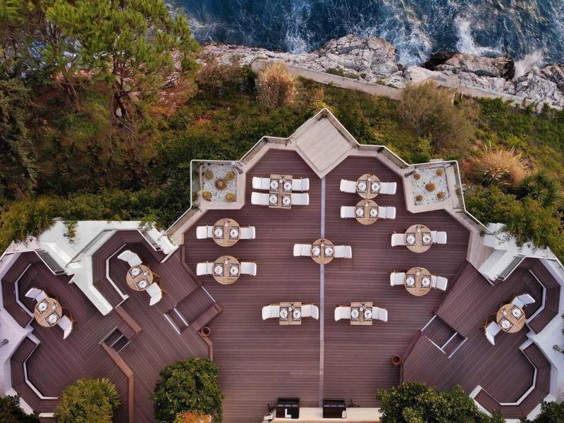 Symmetrische Dachterrasse mit Tischen, Stühlen und Pflanzen vor bewaldetem Hügel und Felsenmeer.