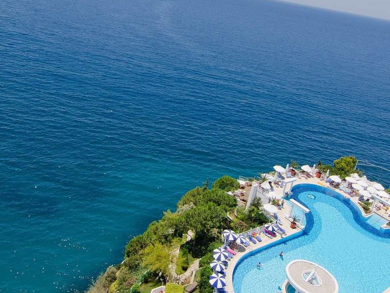 Rundlicher Pool auf Klippe mit Blick auf tiefblaues Meer und Liegestühlen umgeben von Vegetation.