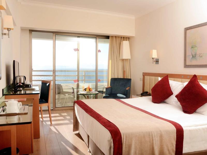 Helles Hotelzimmer mit Doppelbett, roter Bettdecke, Schreibtisch und Balkon mit Meerblick.