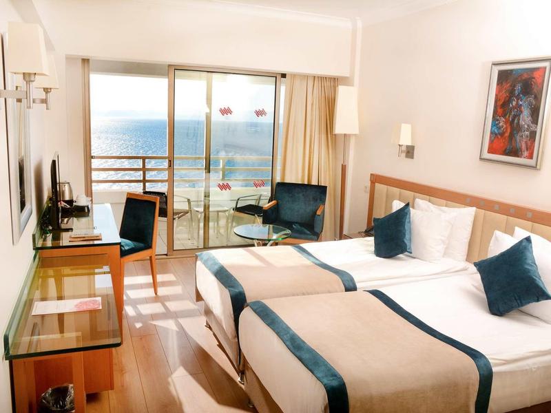 Helles Hotelzimmer mit zwei Betten, Fernseher, Stuhl, Tisch und Balkon mit Meerblick.