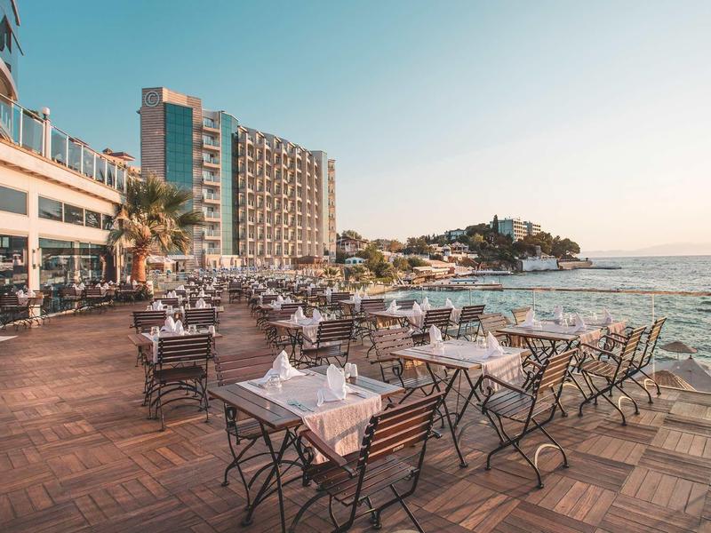 Terrasse mit Tischen und Stühlen am Meer, moderner Hotelbau und bewachsene Hügel im Hintergrund.