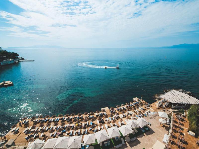 Strand mit Liegestühlen und Sonnenschirmen an klarem, blauem Meer unter bewölktem Himmel.