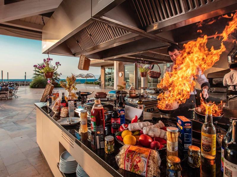 Offene Grillküche mit großer Flamme, vielen Gewürzen, Früchten und Blick aufs Meer.