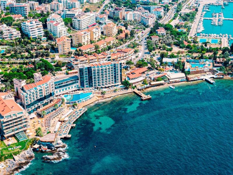 Luftaufnahme von Küstenstadt mit blauer Bucht, Hafen, Gebäuden und grünen Bereichen am Wasser.