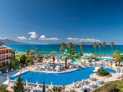 Großes Hotel mit zwei Gebäuden, großem Pool, Palmen und Blick auf das blaue Meer bei klarem Himmel.