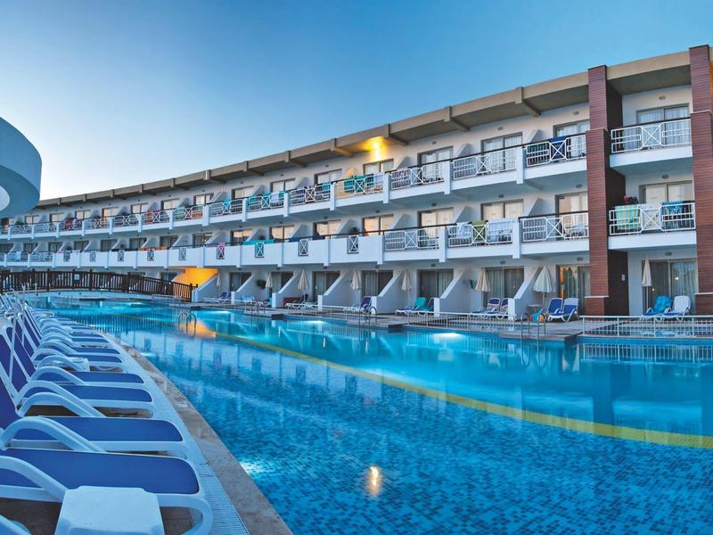 Moderner Hotelkomplex mit langem Pool, blauen Liegestühlen und mehreren Balkonen unter klarem Himmel.