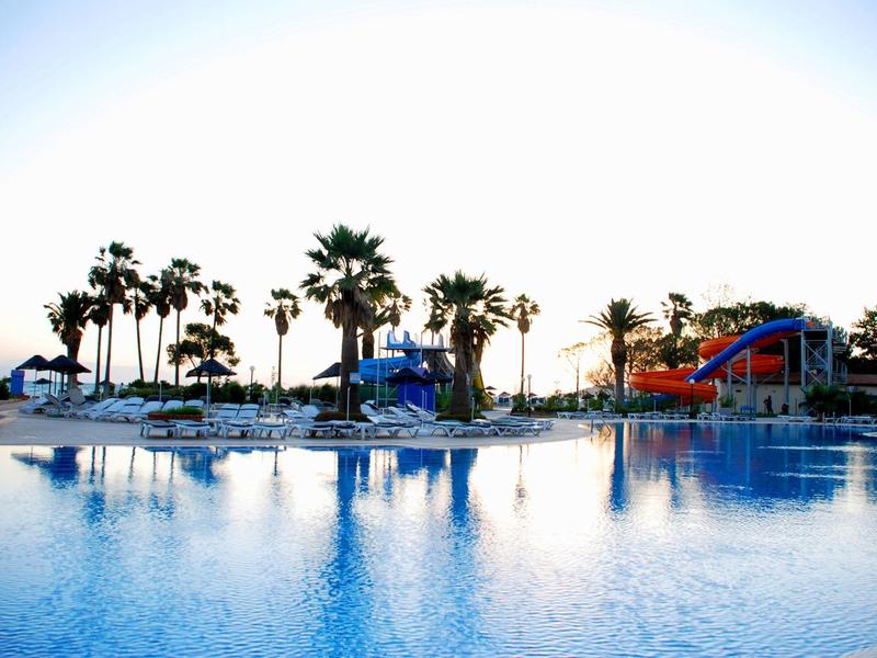 Großes, ruhiges Schwimmbecken mit glattem Wasser, Palmen und Wasserrutsche im Hintergrund bei Sonnenuntergang.