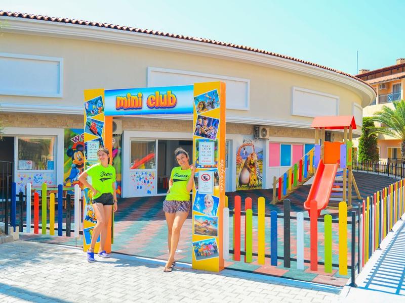 Kinderclub mit buntem Zaun, Rutsche und fröhlichen Betreuerinnen in grellgelben Shirts.