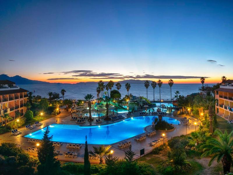 Hotelanlage mit beleuchtetem Pool, Palmen und Gebäuden bei sonnigem Sonnenuntergang.