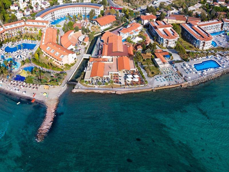 Luftaufnahme eines Küstenresorts mit roten Dächern, mehreren Pools und klarem blauem Meer.