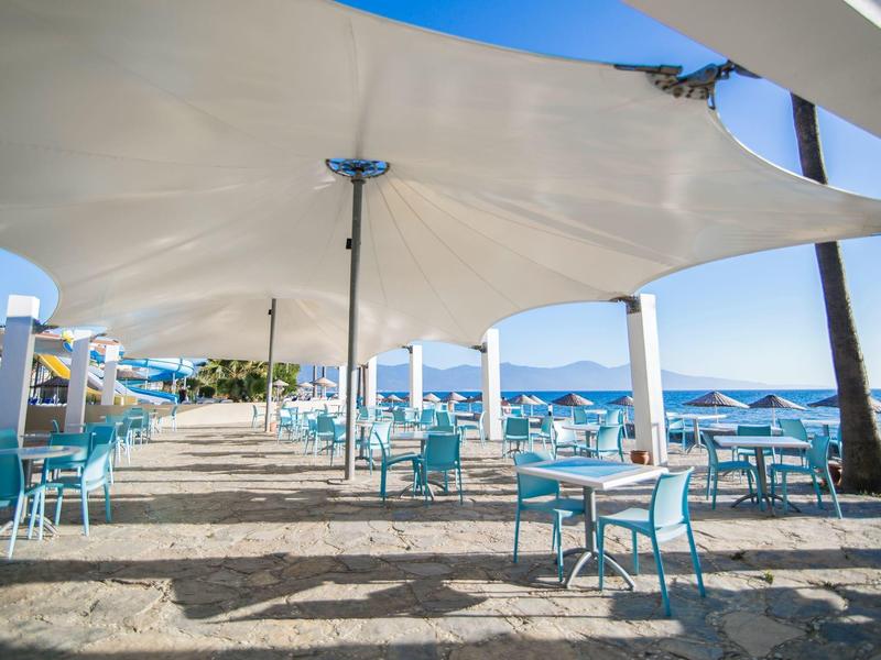 Freier Strandbereich mit blauen Stühlen und weißen Tischen unter hellem Sonnensegel am Meer.